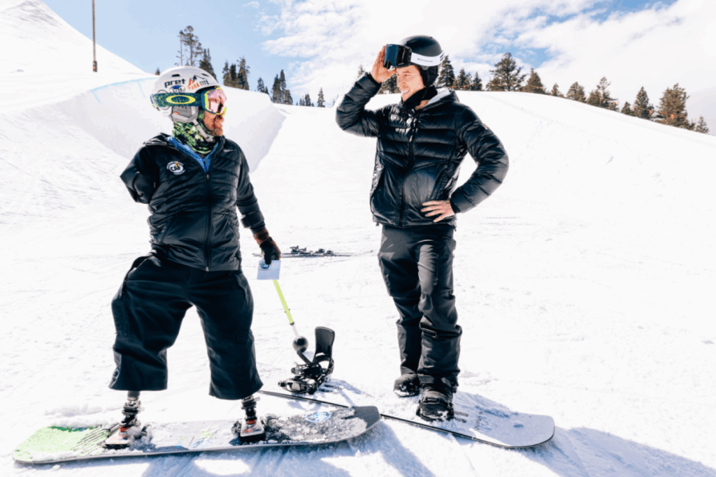 Two snowboarders, one with prosthetic legs, pause on a sunny slope to talk during a winter sports session.