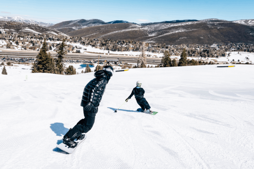 Two snowboarders carve down a wide open mountain run with snow-covered hills and a highway in the background.