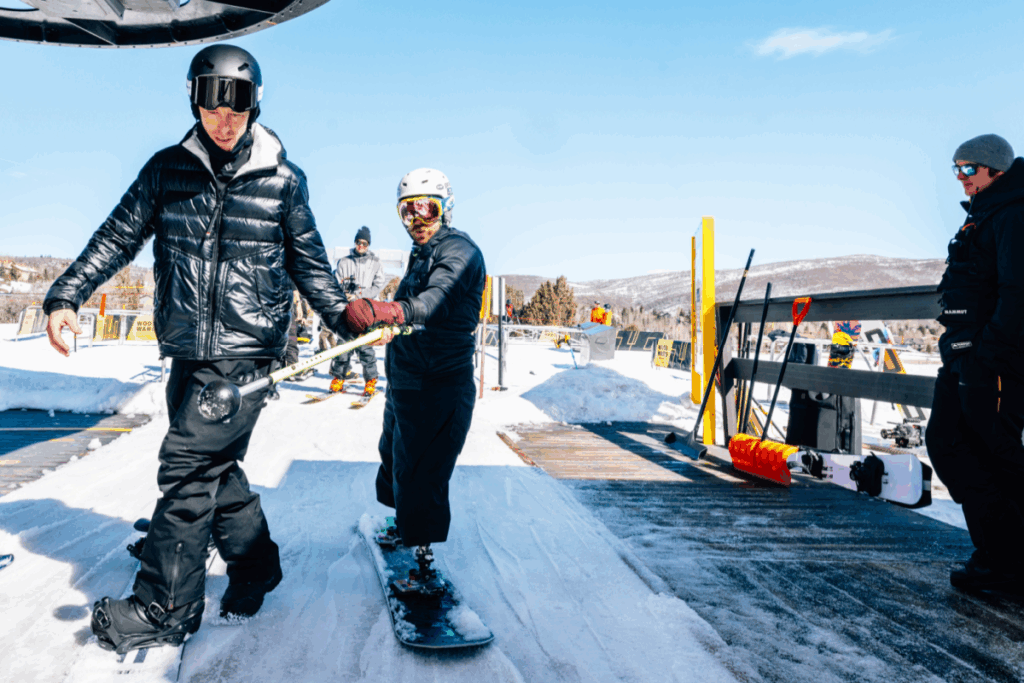 Two snowboarders prepare to board a ski lift on a clear day, one offering support to the other who uses prosthetic legs.