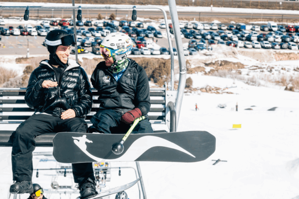 Two snowboarders wearing helmets and winter gear share a laugh while riding a ski lift over a snowy mountain landscape.