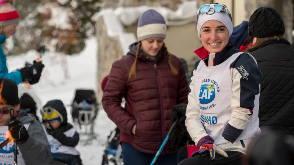 People gathered outdoors in winter clothing, one in the foreground smiling and wearing a white vest with logos.