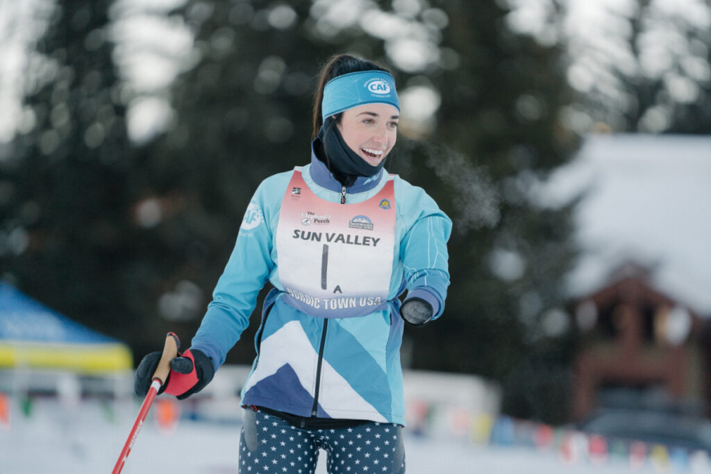 A skier wearing a blue and white outfit and a bib with "SUN VALLEY" on it, outdoors in a snowy forest.