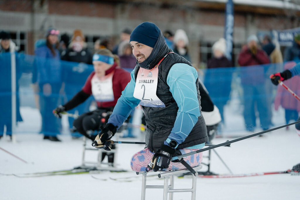 Person in a sit-ski race, wearing a blue head cover and jacket, with spectators in the background.