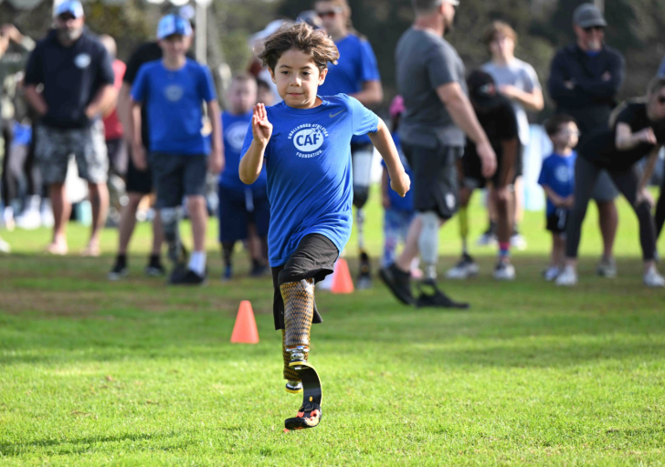 A child wearing a blue CAF shirt and running blade sprints forward on a grassy field, focused and determined, with supportive spectators and other young athletes in the background.