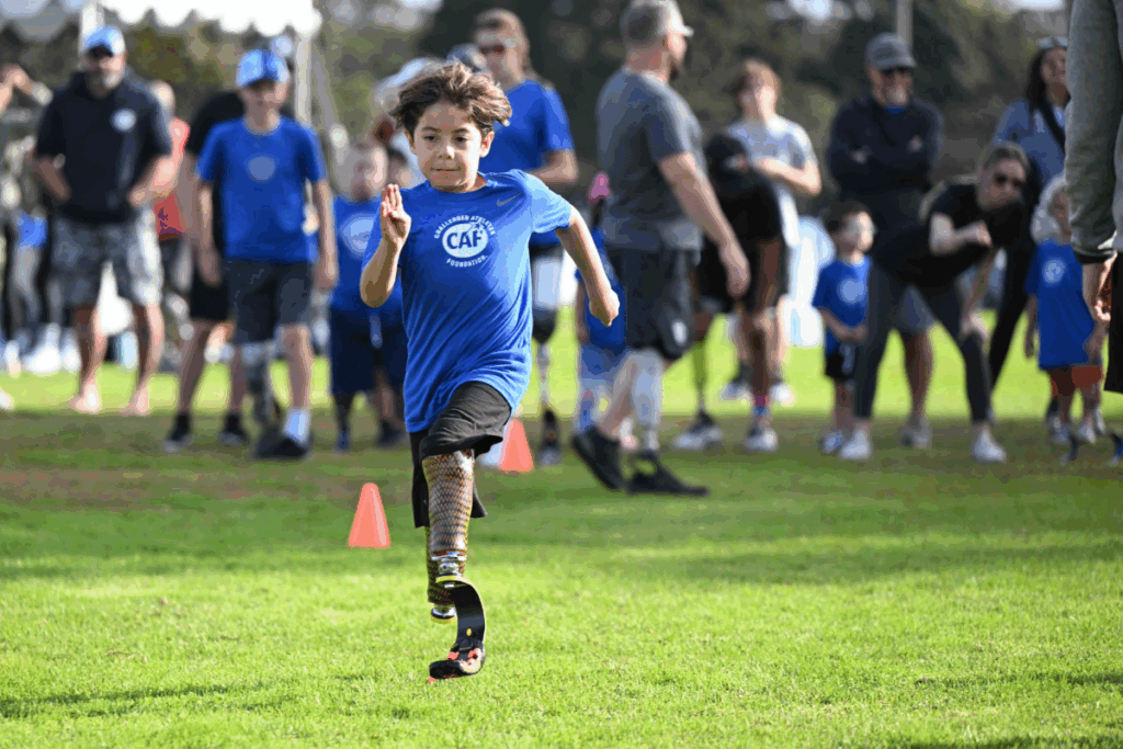 A child wearing a blue CAF shirt and running blade sprints forward on a grassy field, focused and determined, with supportive spectators and other young athletes in the background.