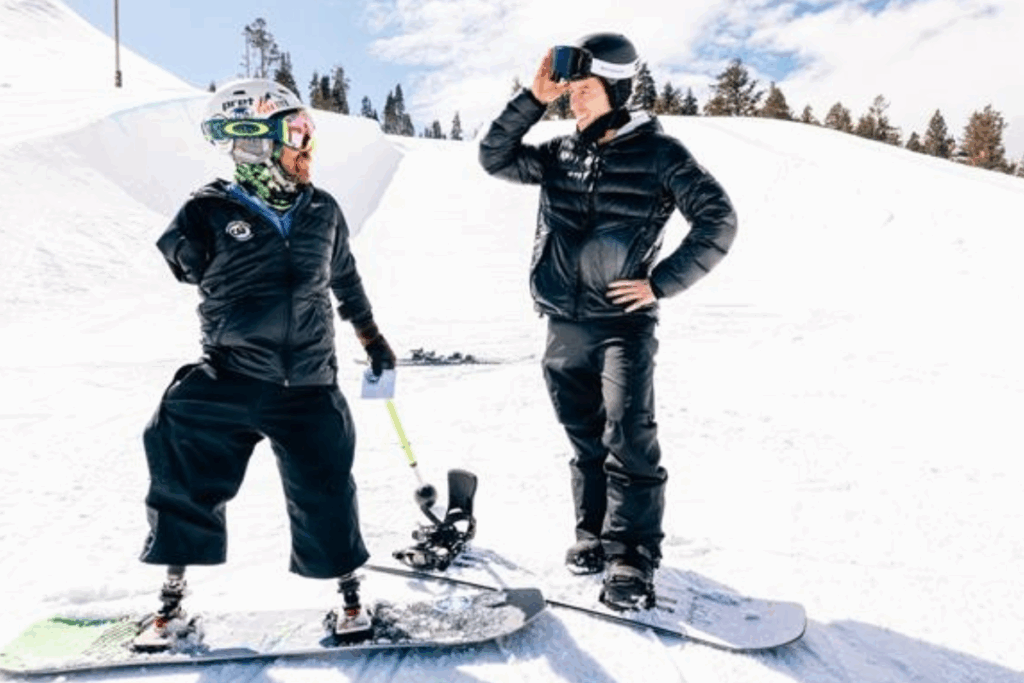 Two snowboarders talk on a snowy mountain slope, one using prosthetic legs and holding adaptive equipment.