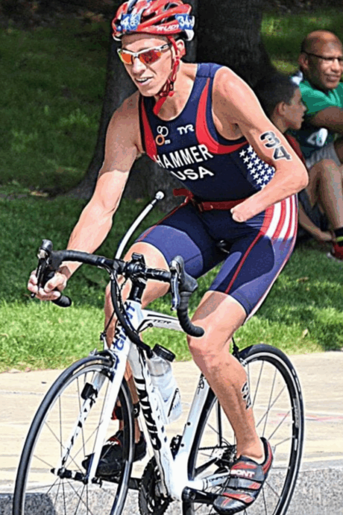 A triathlete rides a road bike during competition, wearing a USA racing suit and helmet.