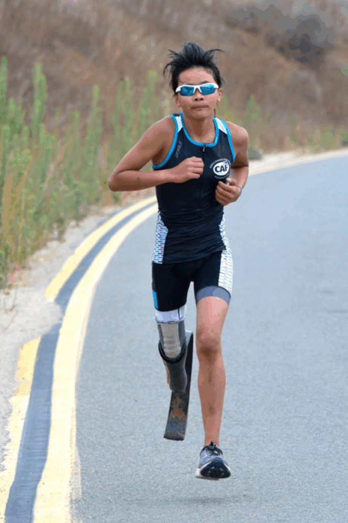 A young runner with a prosthetic leg runs on a paved road, wearing a CAF triathlon suit and sunglasses.