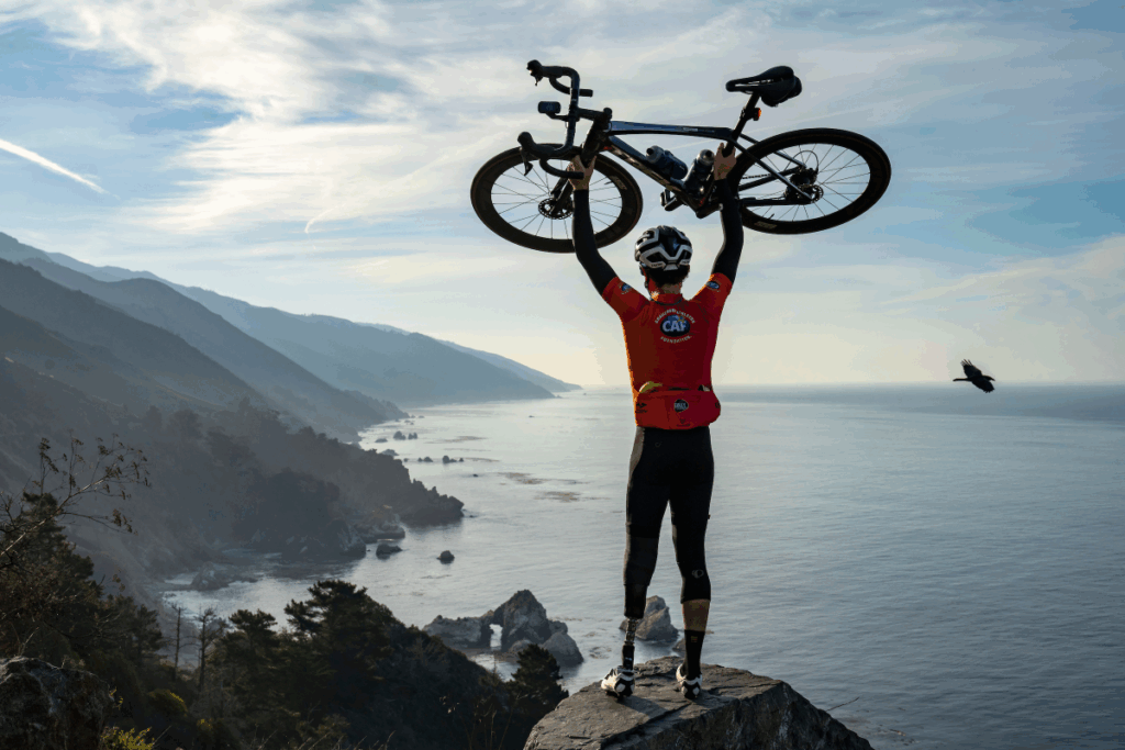 Cyclist wearing a CAF jersey holds their bike overhead on a cliff overlooking the Pacific coastline during Million Dollar Challenge.