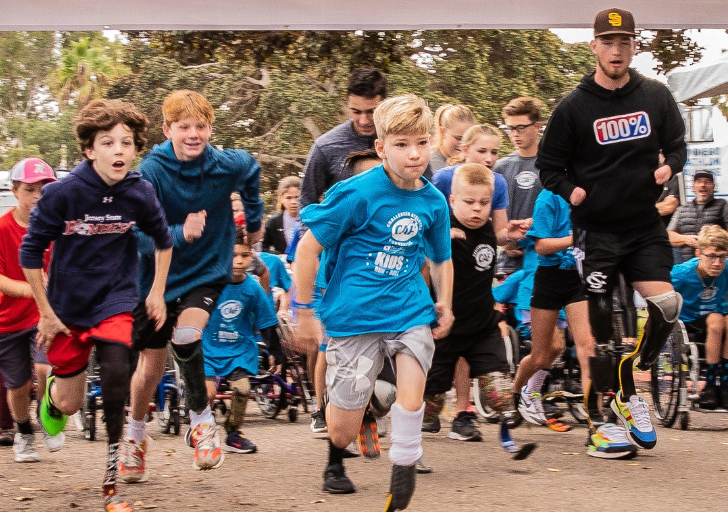 A large group of children in blue CAF shirts, alongside peers of all abilities, sprint forward at the start of a kids’ run, full of energy and determination.
