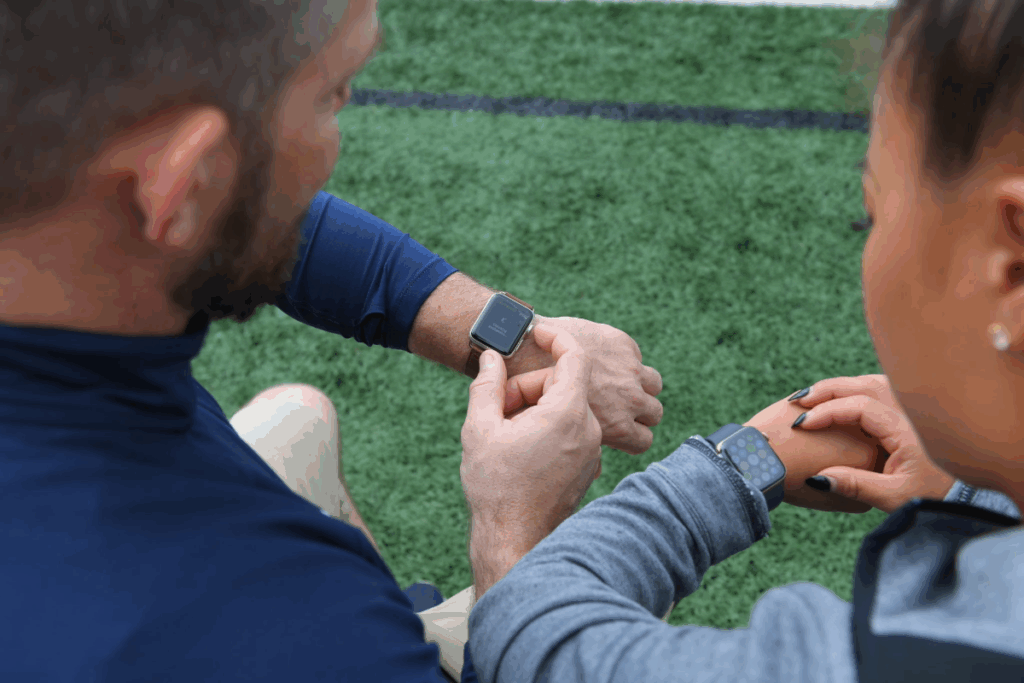 A coach and an athlete compare data on their smartwatches while sitting on a turf field, highlighting technology in training.