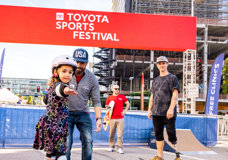 A young girl wearing a helmet practices skateboarding with support from an instructor under a red Toyota Sports Festival banner.