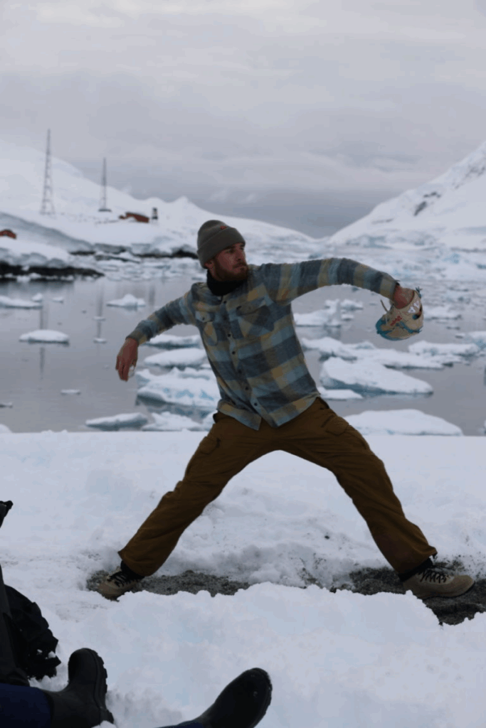 An athlete in a gray-and-blue plaid jacket and beanie throws a baseball on Antarctic snow, framed by icy water and glaciers in the distance.