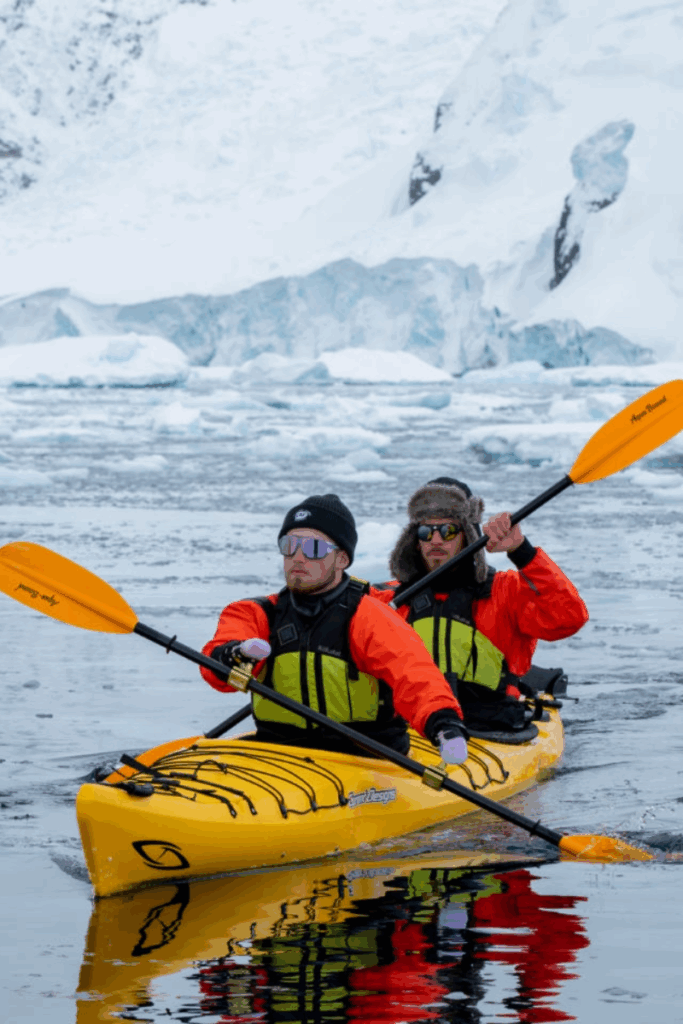 Two athletes paddle a bright yellow tandem kayak through icy Antarctic waters, surrounded by snow-covered mountains and glaciers.