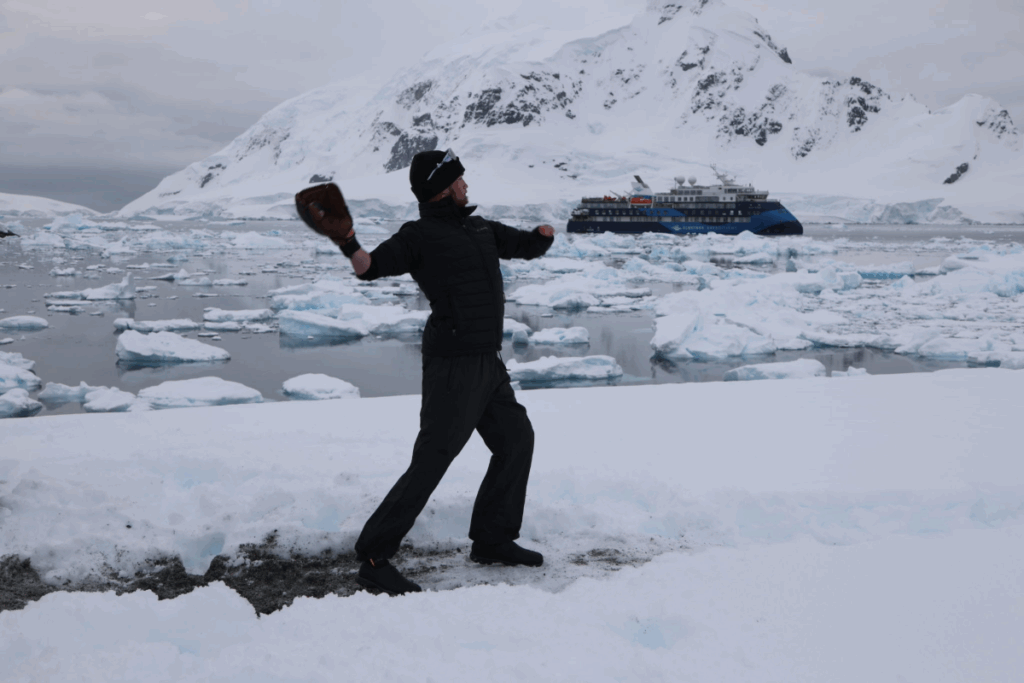 An athlete in dark winter gear winds up to throw a baseball on the snowy Antarctic shoreline, with icebergs and a ship in the background.
