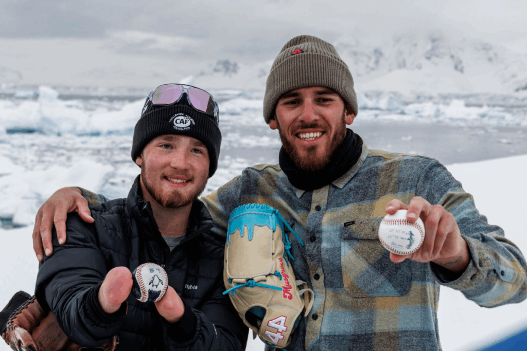 Two smiling men in cold-weather gear stand side by side in Antarctica, holding signed baseballs and a glove, with icy water and mountains behind them.