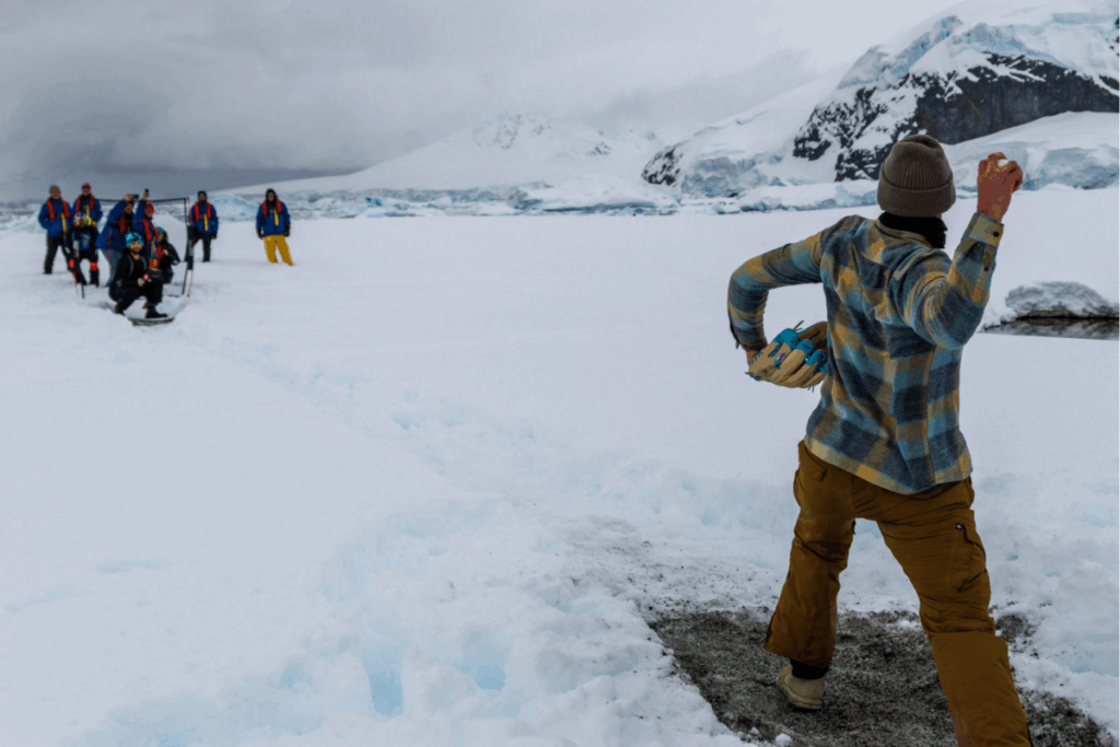 An athlete in a plaid jacket and brown pants throws a baseball on a makeshift snowy field, with teammates watching in the distance against icy peaks.