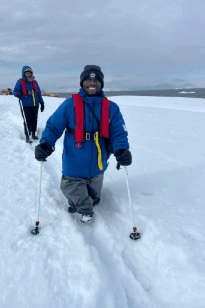 A smiling athlete with a prosthetic leg hikes through deep Antarctic snow using trekking poles, wearing a blue jacket and red life vest.