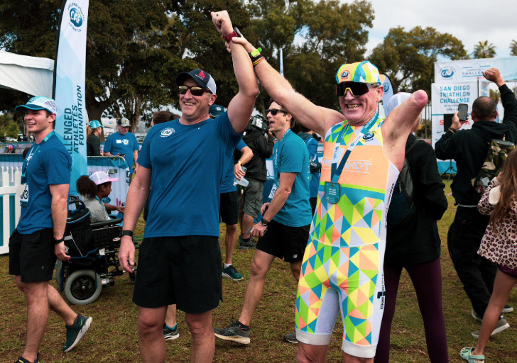 An athlete with a prosthetic arm raises both fists in celebration at the finish line of the San Diego Triathlon Challenge, smiling alongside a supporter holding up his arm in victory.