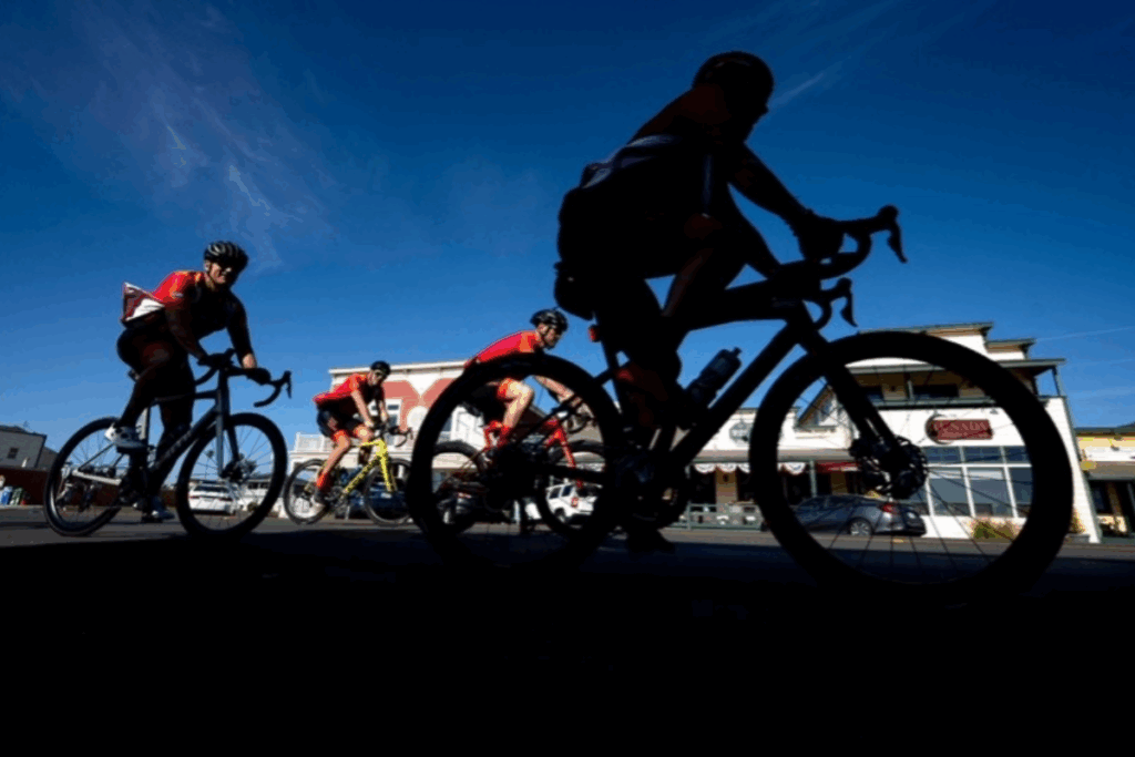 A group of cyclists ride together on a sunny street, silhouetted against a clear blue sky.