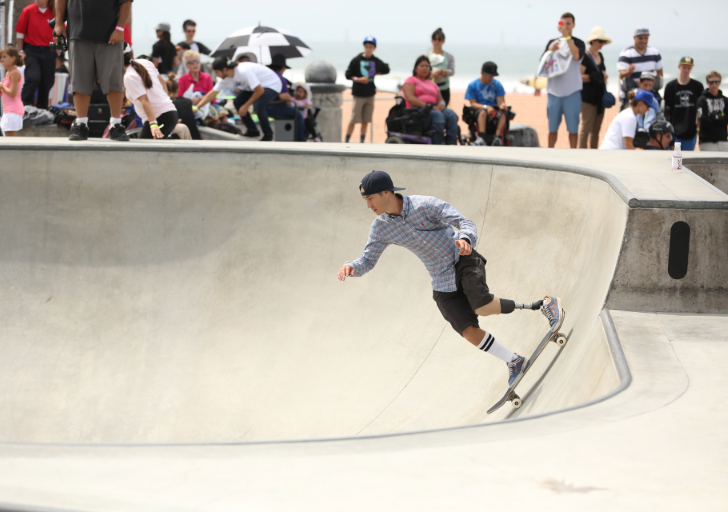 A skateboarder with a prosthetic leg rides along the curved wall of a concrete skatepark bowl, wearing a plaid shirt, black shorts, and a backwards baseball cap.
