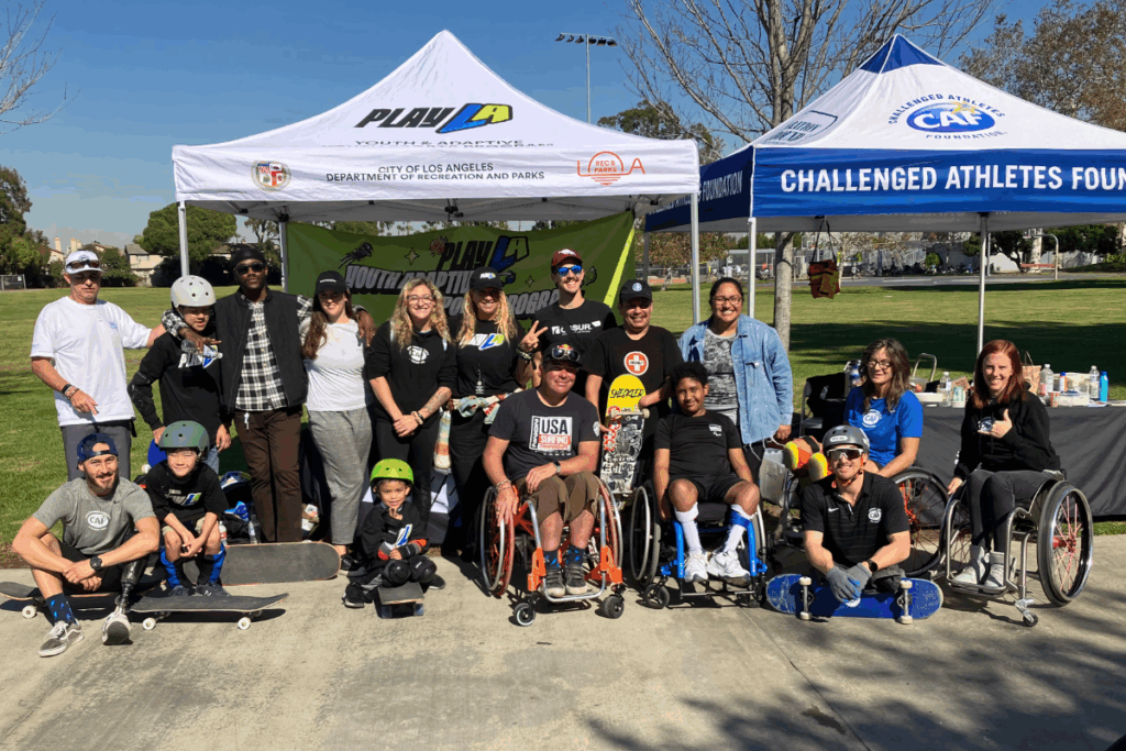 A large group of athletes, coaches, and supporters gather for a group photo under tents with banners for PlayLA and the Challenged Athletes Foundation. Some athletes are in wheelchairs, while others hold skateboards.