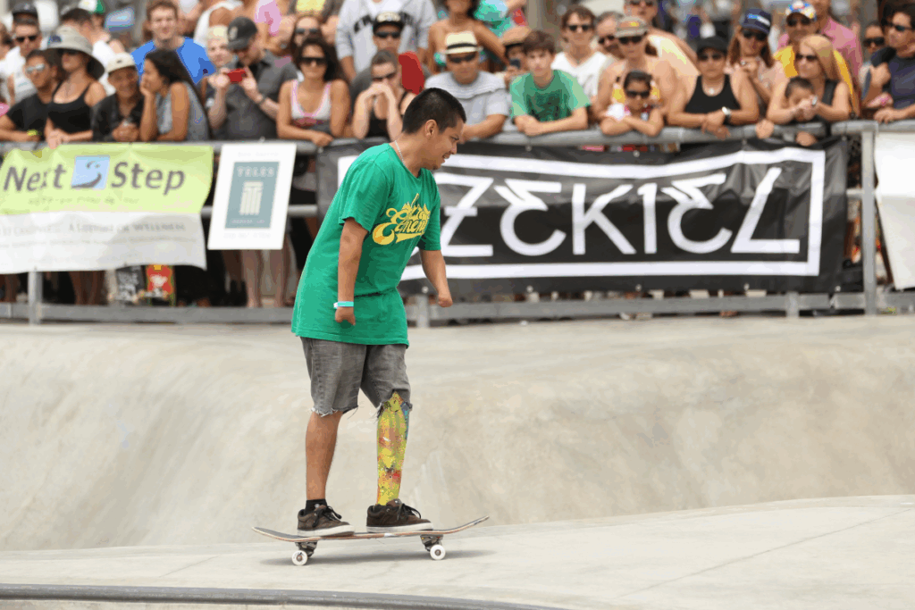 A skateboarder with a prosthetic leg and limb difference rides confidently across the skatepark bowl while a crowd of spectators watches from behind the fence.