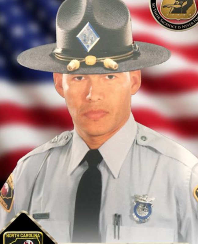 A formal portrait of a uniformed North Carolina state trooper in front of a blurred American flag backdrop. He wears a gray shirt, tie, and campaign hat with a badge, symbolizing service and honor.