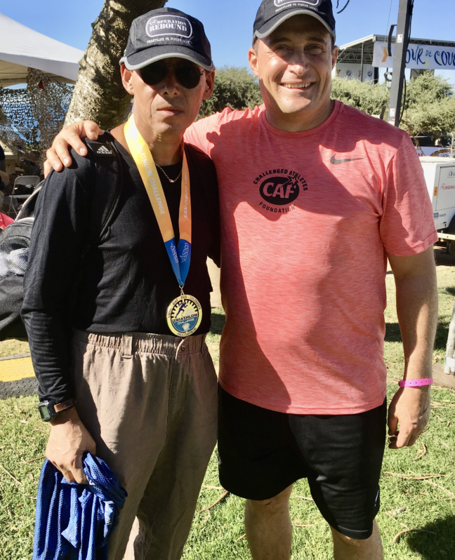 Two men stand smiling together outdoors at a Challenged Athletes Foundation event. The man on the left wears sunglasses, a hat, and a medal around his neck, while the man on the right wears a CAF shirt and cap.