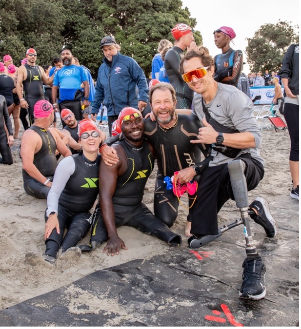 Three triathletes, including an athlete with a prosthetic leg, smile and pose on the beach surrounded by other swimmers.