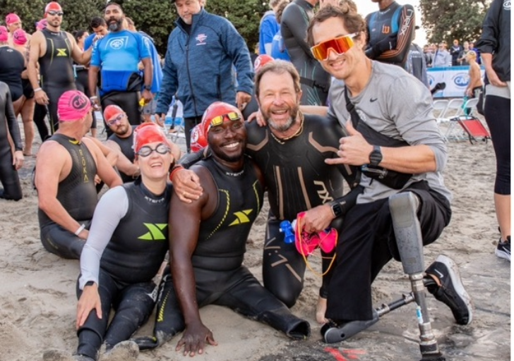Group of triathletes, including an athlete with a prosthetic leg, smile and pose together on the beach before a swim start.