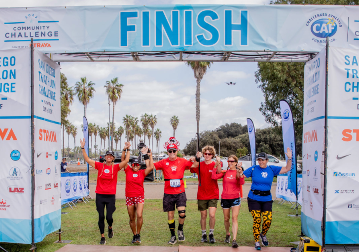 Six athletes in matching red shirts and one in blue raise their hands together in victory as they cross the finish line at the CAF Community Challenge, with palm trees and banners in the background.