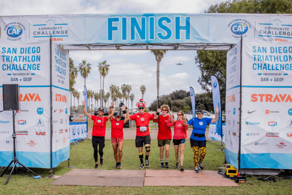 Six athletes in matching red shirts and one in blue raise their hands together in victory as they cross the finish line at the CAF Community Challenge, with palm trees and banners in the background.