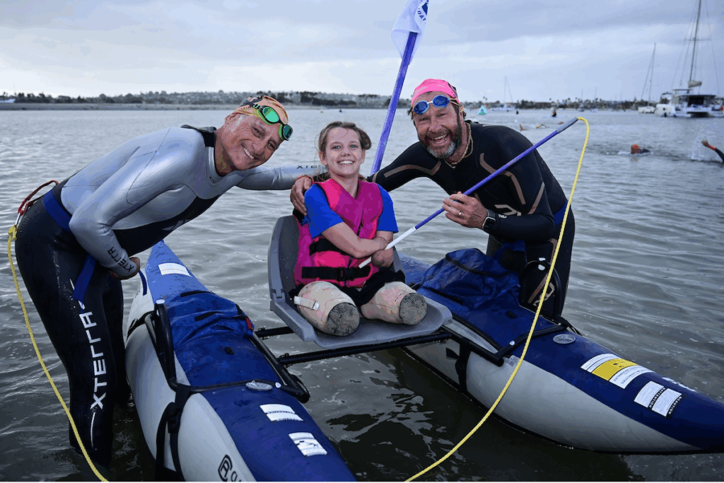 Two wetsuit swimmers in goggles smile beside a young adaptive athlete seated on a raft-like watercraft, holding a flag as they prepare for the swim event.