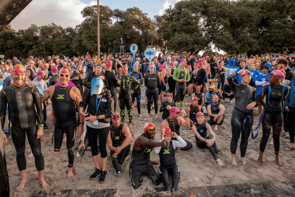 Dozens of wetsuit swimmers wearing colorful swim caps gather on the sand, waiting for the race start, with some adjusting goggles and others cheering.