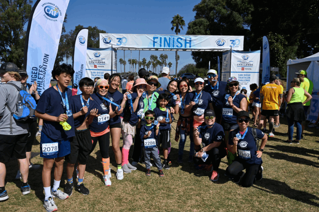 A large group of participants wearing CAF shirts and medals posing proudly under the finish line arch at the San Diego Triathlon Challenge.