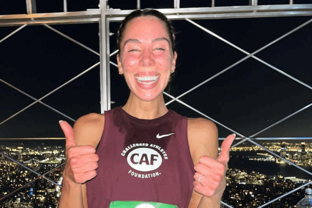 A joyful female athlete in a Challenged Athletes Foundation singlet, flushed from exertion, smiling broadly and giving two thumbs up at the top of the Empire State Building against the New York City night skyline.