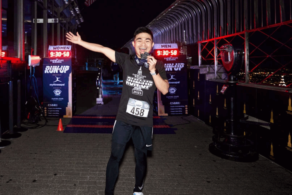 A male runner celebrating at the Empire State Building observatory after completing the Run-Up race, smiling, holding his medal in his mouth, and posing triumphantly with the city lights glowing in the background.