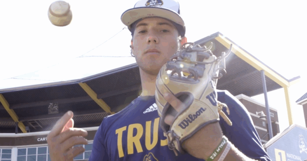A baseball player wearing a purple “TRUST” shirt and glove tosses a ball into the air outside a stadium.