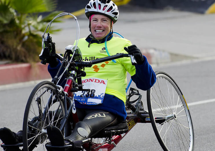 An athlete with a physical disability smiles while racing a handcycle on a paved road, wearing a helmet and bright race shirt during a long-distance endurance event.