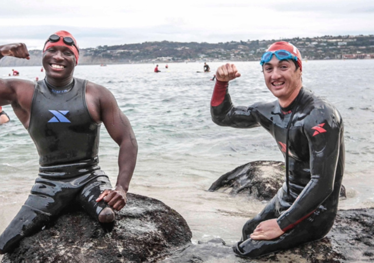 Two athletes, Rudy Garcia-Tolson and Roderick Sewell, in wetsuits and swim caps smiling and flexing their arms while sitting on rocks at the edge of the ocean after a swim.