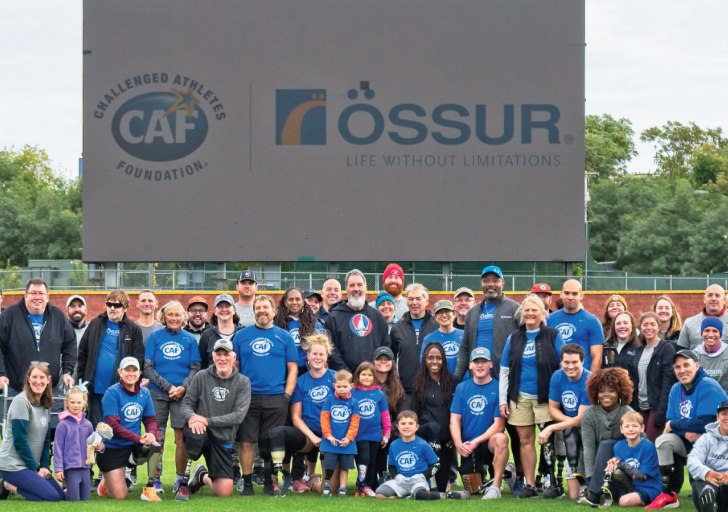 A large group of athletes, families, and supporters pose together on a sports field in front of a giant screen displaying the Challenged Athletes Foundation (CAF) and Össur logos.