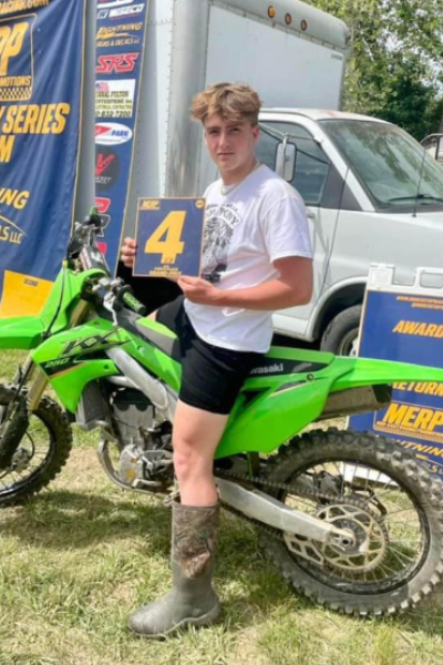 A motocross rider in a white shirt and shorts stands next to a green dirt bike, holding a “#4” award plaque, with event banners in the background.