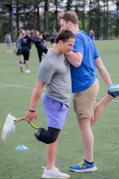Two athletes stretch together on a turf field before training, one using a running blade prosthetic while balancing with a partner.
