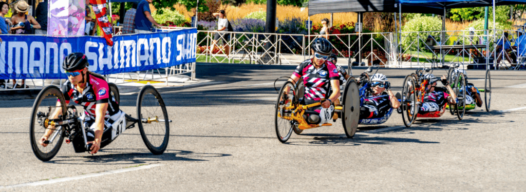 A group of athletes with physical disabilities racing in handcycles on a city street, wearing matching team jerseys and helmets. Spectators line the course behind barricades, cheering as the athletes push forward in the competition.