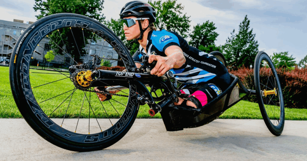 Close-up of an athlete with a physical disability in a racing handcycle, wearing a helmet, sunglasses, and a black-and-blue team jersey. The athlete leans forward with focus, gripping the hand pedals tightly.