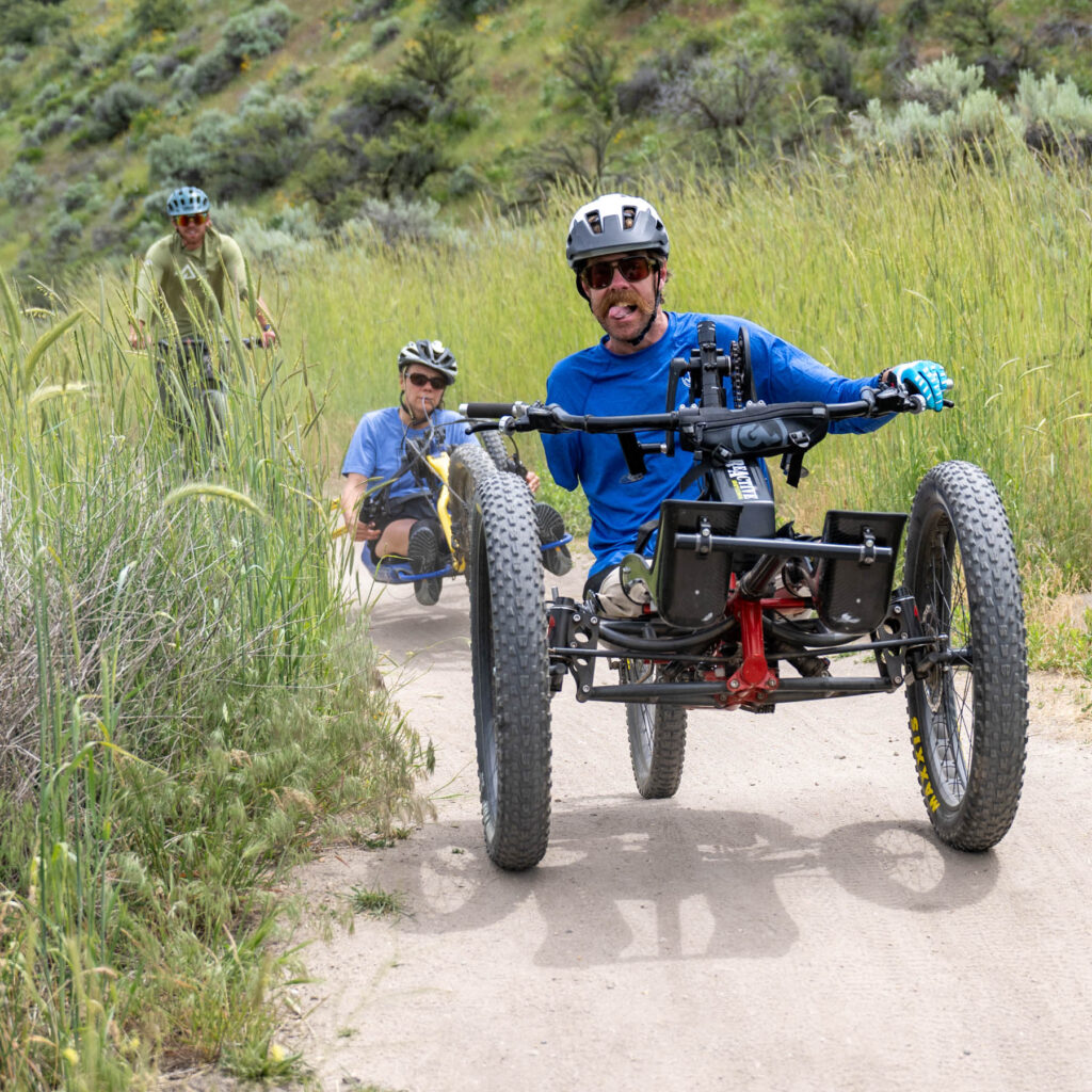 Two people riding adaptive mountain bikes and one person on an upright cycle on a dirt trail. The person in the front has three limb differences and is sticking out their tongue.