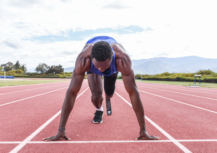 A sprinter with a running blade, Derek Loccident, crouches at the starting line on a red track, focused before a sprint.