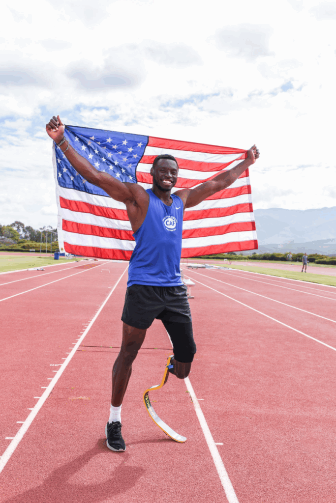 An athlete with a running blade, Derek Loccident, holds an American flag overhead and smiles on a track.