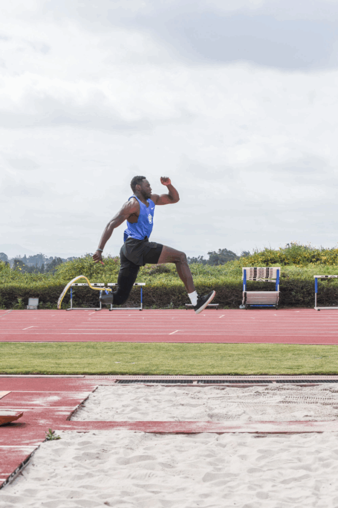 An athlete with a running blade, Derek Loccident, leaps mid-air during long jump practice on a red track.